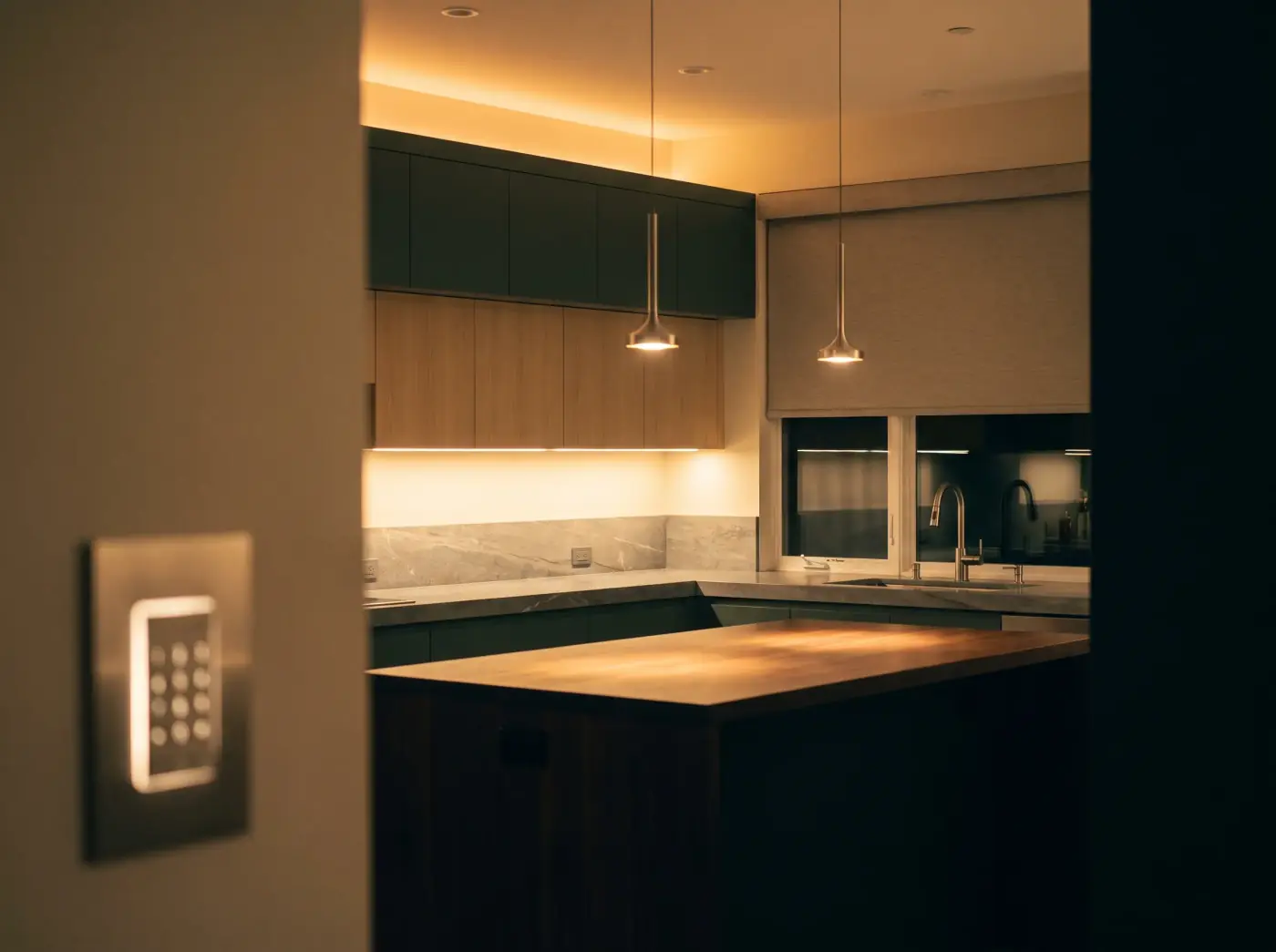 A kitchen at night lit by warm under-cabinet and cove lighting, with a lowered motorized shade and a backlit wall keypad in the foreground.